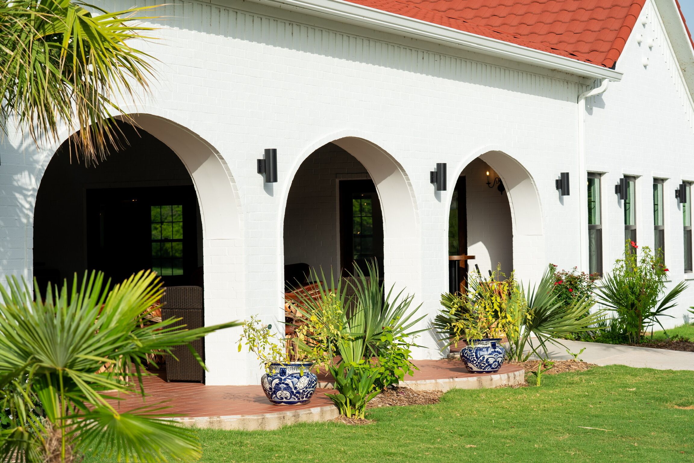 A white building with a red roof, featuring three arched doorways and a well-maintained lawn with potted plants.