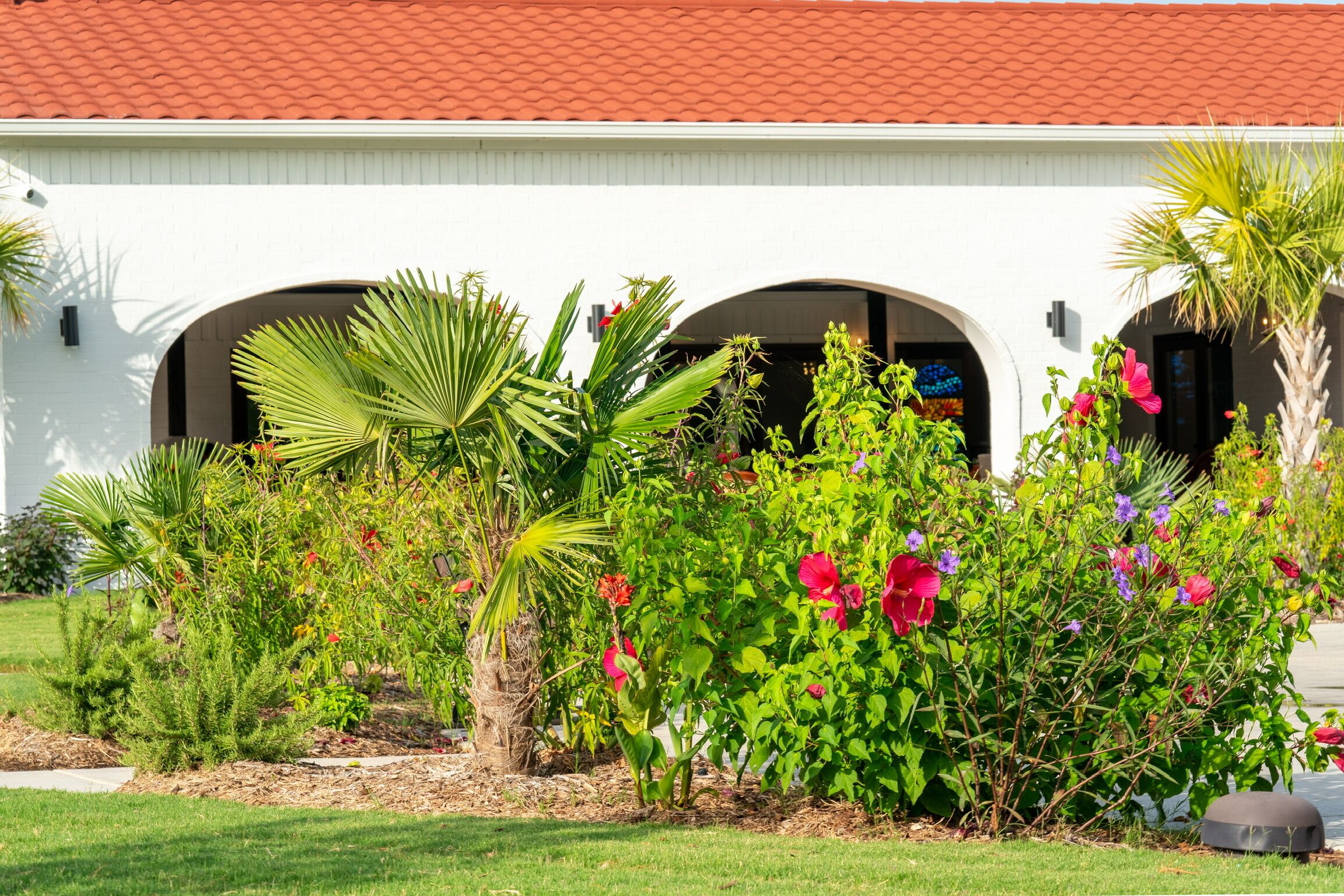 A white building with a red-tiled roof and arched doorways, surrounded by lush greenery and flowering plants.
