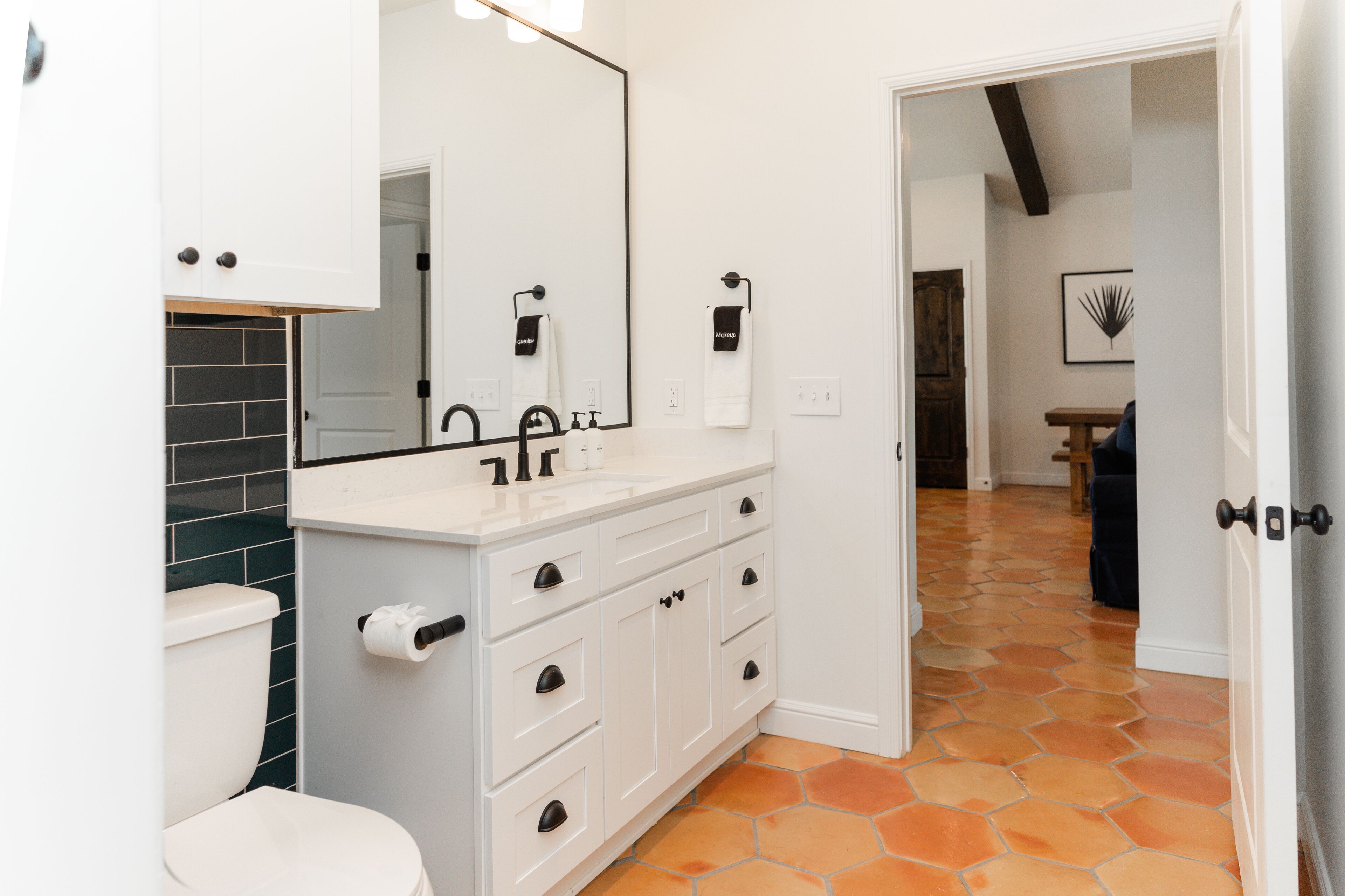 Um banheiro com pia branca, torneira preta e um espelho grande. Há um vaso sanitário com tampa branca e um revestimento de azulejos pretos. O chão possui azulejos hexagonais laranja.