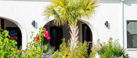 Un edificio blanco con tejado rojo, arcos en las entradas y un pequeño jardín con una palmera y flores de colores.