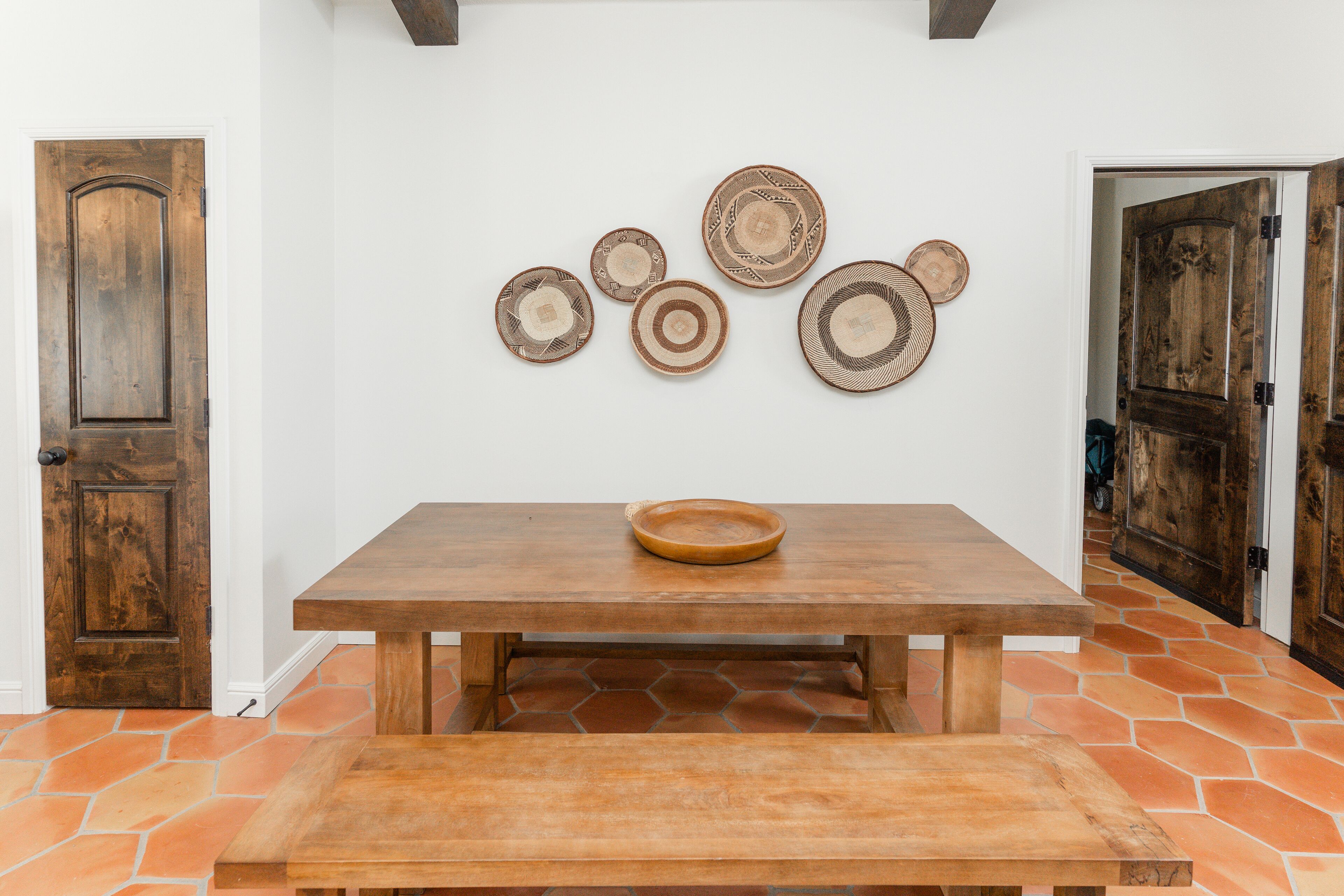 Una mesa de comedor de madera con un tazón encima, rodeada de baldosas hexagonales y una pared con platos decorativos.