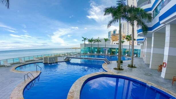 A pool area with a view of the ocean, palm trees, and a building with a blue and white facade. - 2P1-AL3 APARTAMENTO DE 2 ALC CON WIFI (Cartagena)