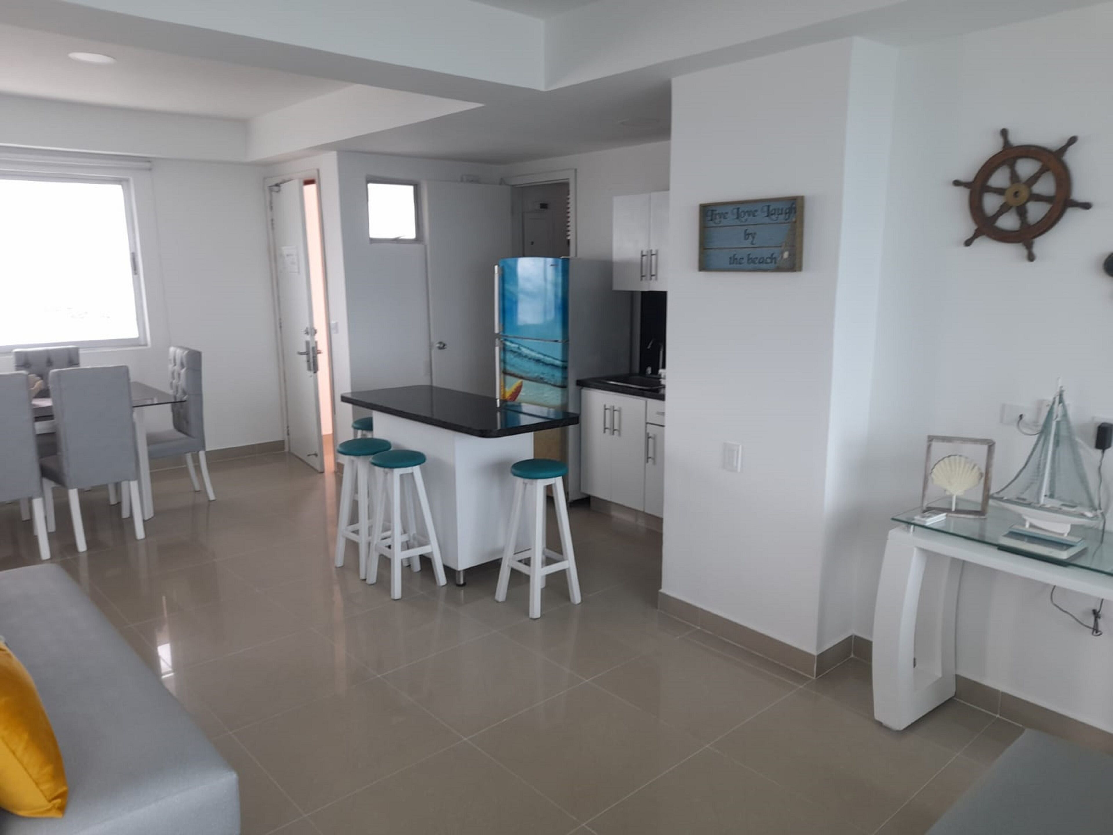 Cuina moderna amb armaris blancs, tauleig negre i una petita illa amb tamborets. Hi ha una zona de dinar amb taula i cadires blanques, i una illa de cuina amb tamborets. Una roda de vaixell decorativa i un cartell emmarcat hi ha a la paret.