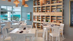 A restaurant with white tables and chairs, a wooden bookshelf wall, and large windows overlooking the ocean. - STUDIO 3 AT THE BEST BEACH IN CANCUN (Cancún)