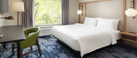 A hotel room with a large bed, a desk, a chair, a window with curtains, and a view of trees.