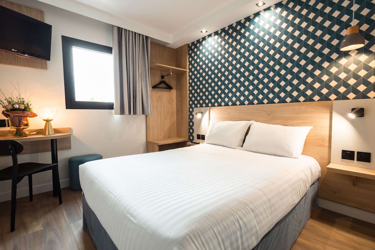 A modern hotel room with a large bed, a desk with a chair, a TV, and a window with curtains.