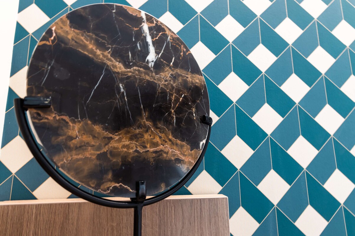 A round mirror with a black marble-like surface, mounted on a wooden stand, against a wall with a geometric tile pattern.