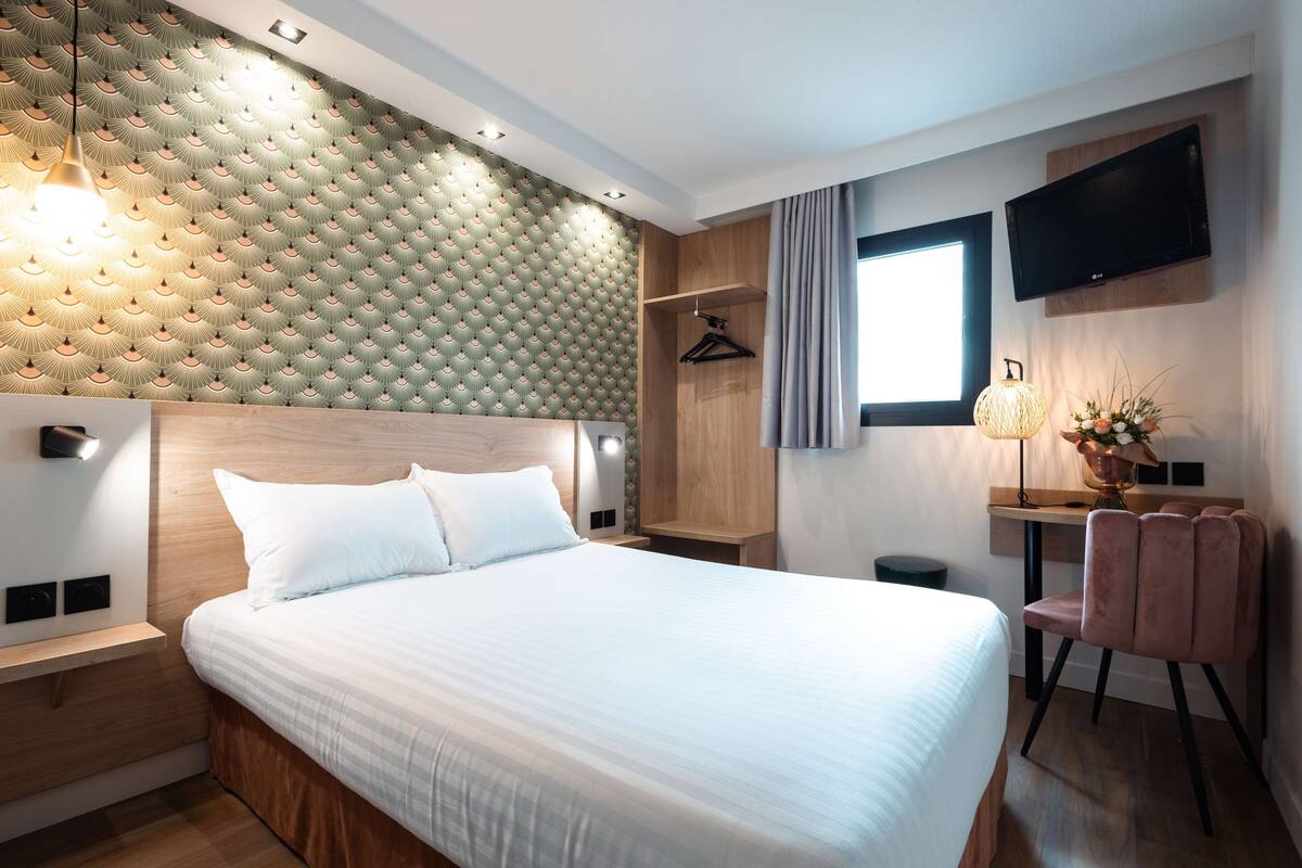 A modern hotel room with a large bed, a wooden headboard, a desk with a chair, a TV mounted on the wall, and a patterned wall.