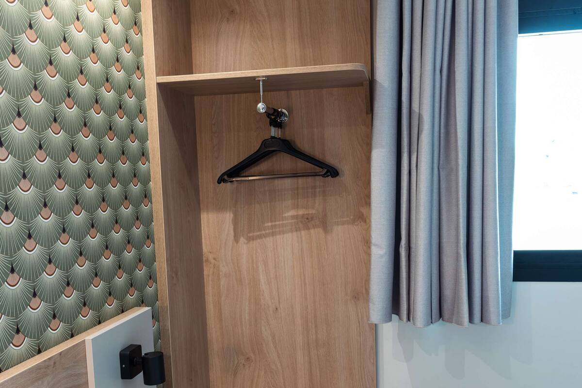 A compact hotel room with a bed, a wooden wardrobe, a small seating area, and a window with curtains.