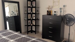 Ein Schlafzimmer mit einem Bett, einer Kommode, einem Spiegel, einem an der Wand befestigten Regal mit Bildern, einem Ventilator und einer Deckenleuchte.