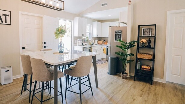 Une cuisine moderne avec des armoires blanches, un comptoir en marbre et un four encastré. On y trouve un coin repas avec une table blanche et quatre chaises. Une grande plante se trouve dans un coin, et une étagère noire ornée d’objets décoratifs est fixée au mur.