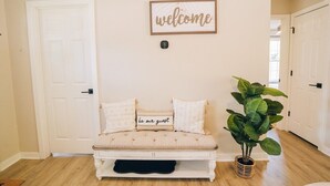 Un banc blanc avec un coussin sur lequel on peut lire « soyez les bienvenus », adossé à un mur beige, avec une pancarte « bienvenue » au-dessus, et un pot de fleurs à droite.