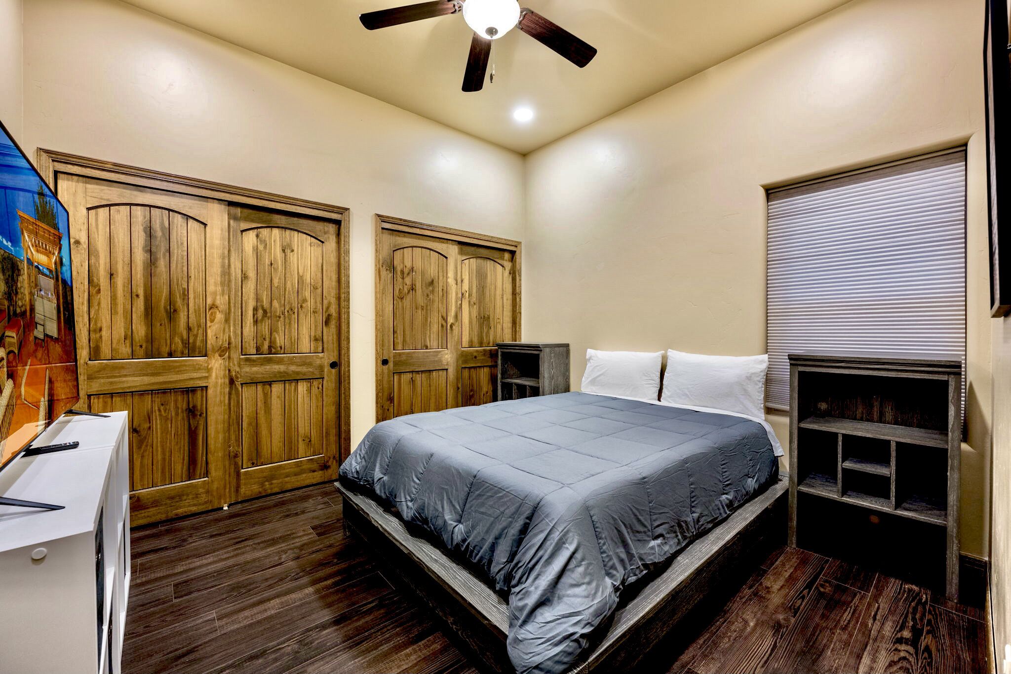 A bedroom with a bed, wooden wardrobe, ceiling fan, and a window with blinds.
