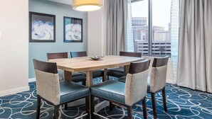 A hotel room with a round table, six chairs, a large window with curtains, and two abstract paintings on the wall.
