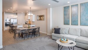 A modern living room with a sofa, a dining area with chairs, a kitchen with cabinets, and a ceiling fan. - Poolside Plunge (St. George)
