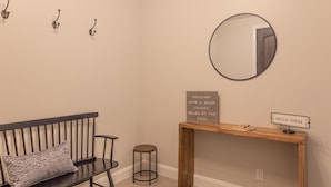 A room with a bench, a round mirror, a small stool, a wooden console table with a sign, and wall-mounted hooks. - Poolside Plunge (St. George)