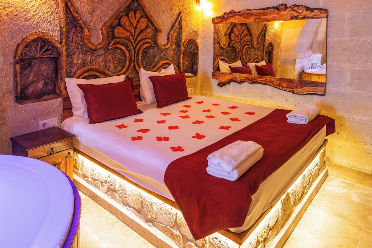 Un dormitorio con cama, almohadas, una mesita de noche, un espejo y una pared con detalles decorativos tallados.