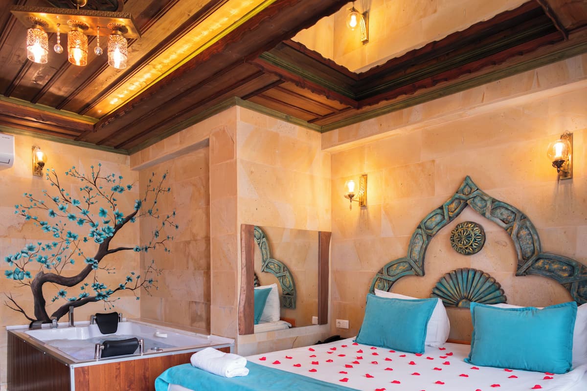 Un dormitorio con pared de piedra, techo de madera, cama con ropa de cama azul y blanca, un lavamanos y un mural decorativo con un árbol.