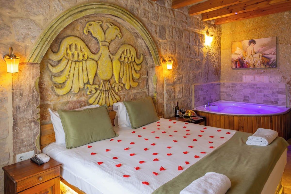 Un dormitorio con pared de piedra, vigas de madera, cama con colcha verde, mesita de noche, lámpara de pared y un cuadro.