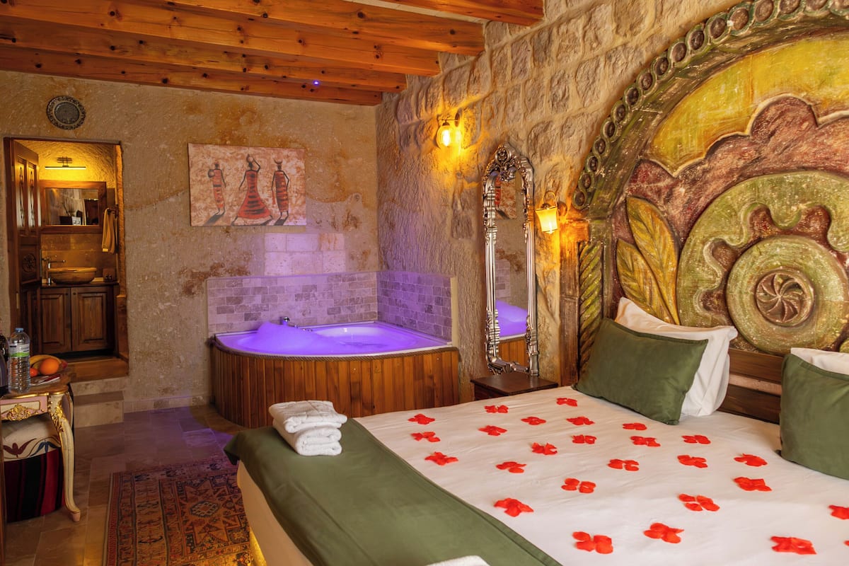 Una habitación con pared de piedra, vigas de madera, cama con ropa de cama verde, bañera y espejo.
