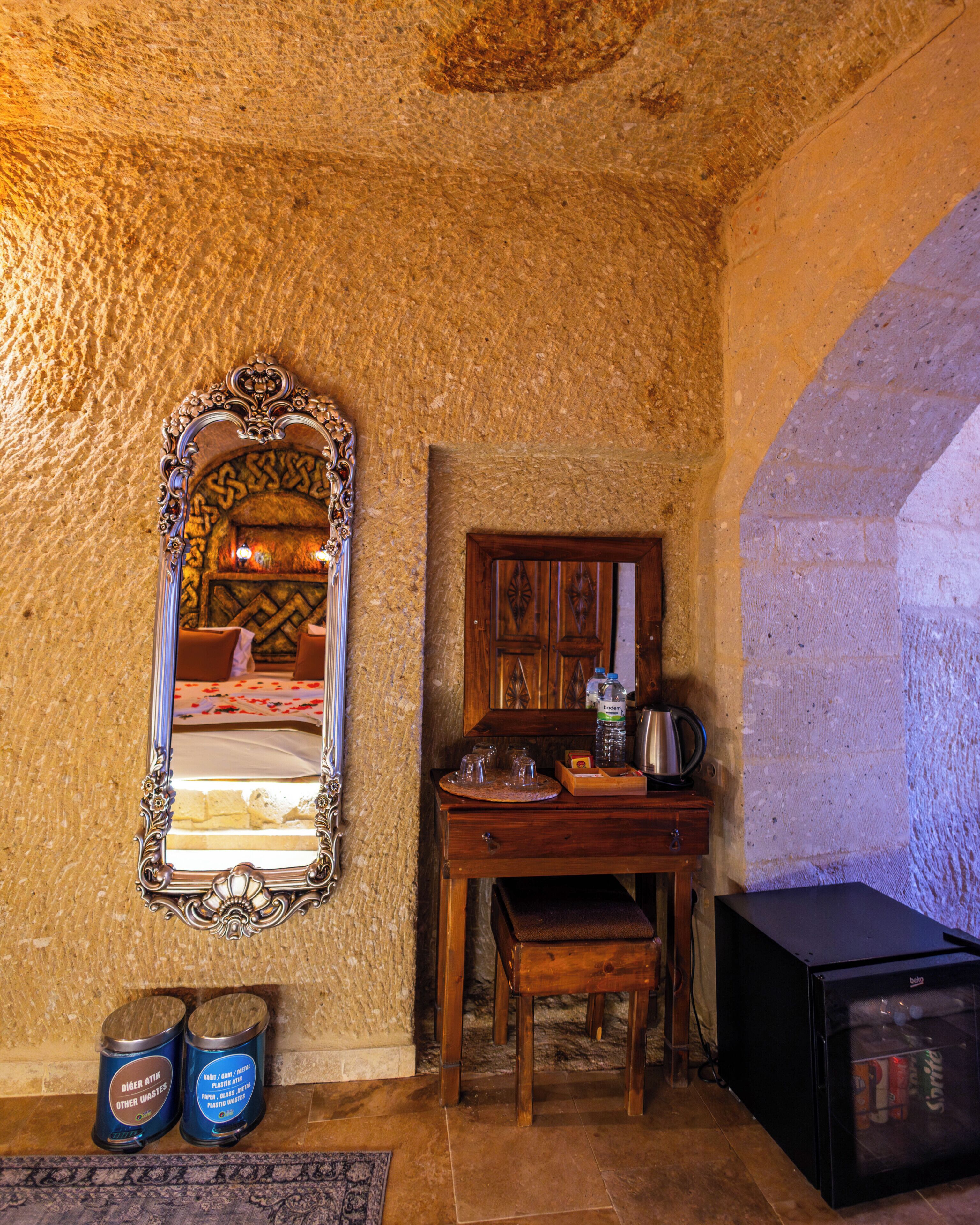 Una habitación rústica con una mesa de madera, un espejo con marco decorativo, una silla, un mueble pequeño y dos cubos de basura etiquetados 'DÖBER ATH' y 'OTROS RESIDUOS'.