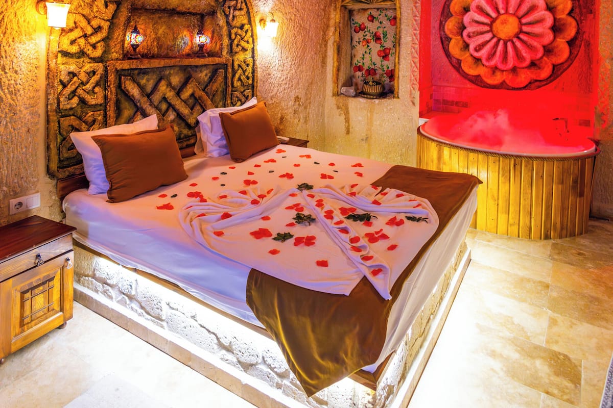 Una habitación con pared de piedra, cama con pétalos de rosa, cabecero de madera y un cuadro de una flor roja.