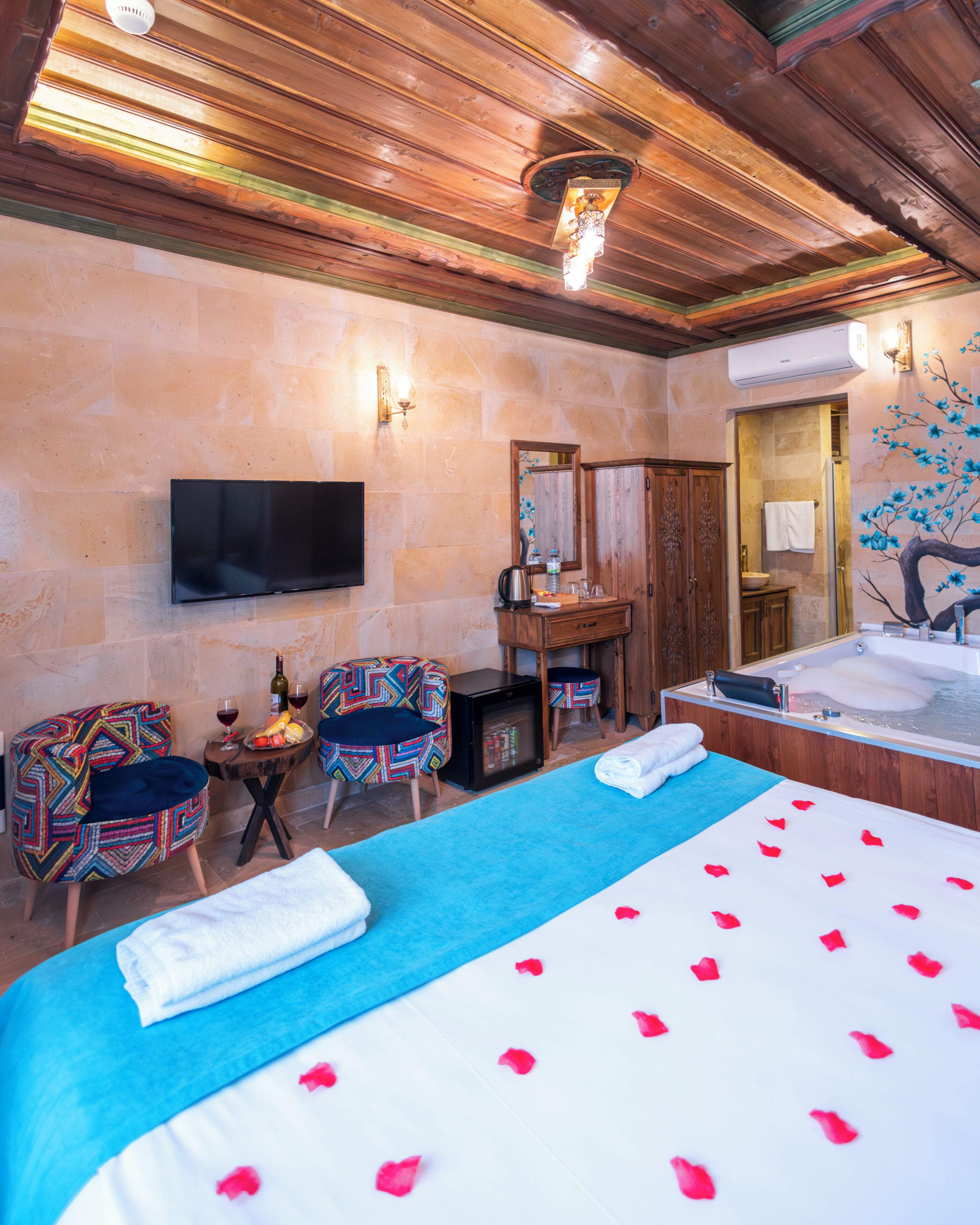 Una habitación de hotel con una cama grande, un televisor, un armario de madera, una mesita con una botella y vasos, y un baño con bañera.