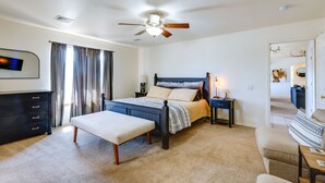 Quarto com cama, um banquinho, mesa de cabeceira, cômoda, ventilador de teto e porta para outro cômodo.
