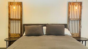 Een bed met een donkerdekkens, twee nachtkastjes met lampen, en een houten hoofdbord met verticale latten.