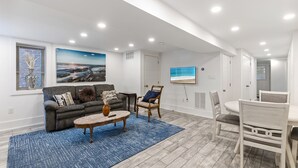 Un vaste espace de vie comprenant un canapé gris, une table basse ronde et un tapis bleu. On y trouve également une table à manger avec des chaises, ainsi qu’un tableau représentant une scène de plage accroché au mur.