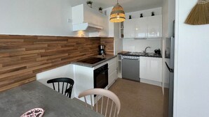 A modern kitchen with white cabinets, a wooden backsplash, a dining area with two chairs, and a hanging light fixture. - Côté Thalasso (Quiberon)