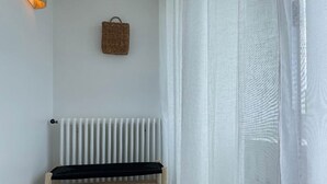 A room with a white radiator, a wooden bench, a woven basket on the wall, and sheer white curtains. - Côté Thalasso (Quiberon)
