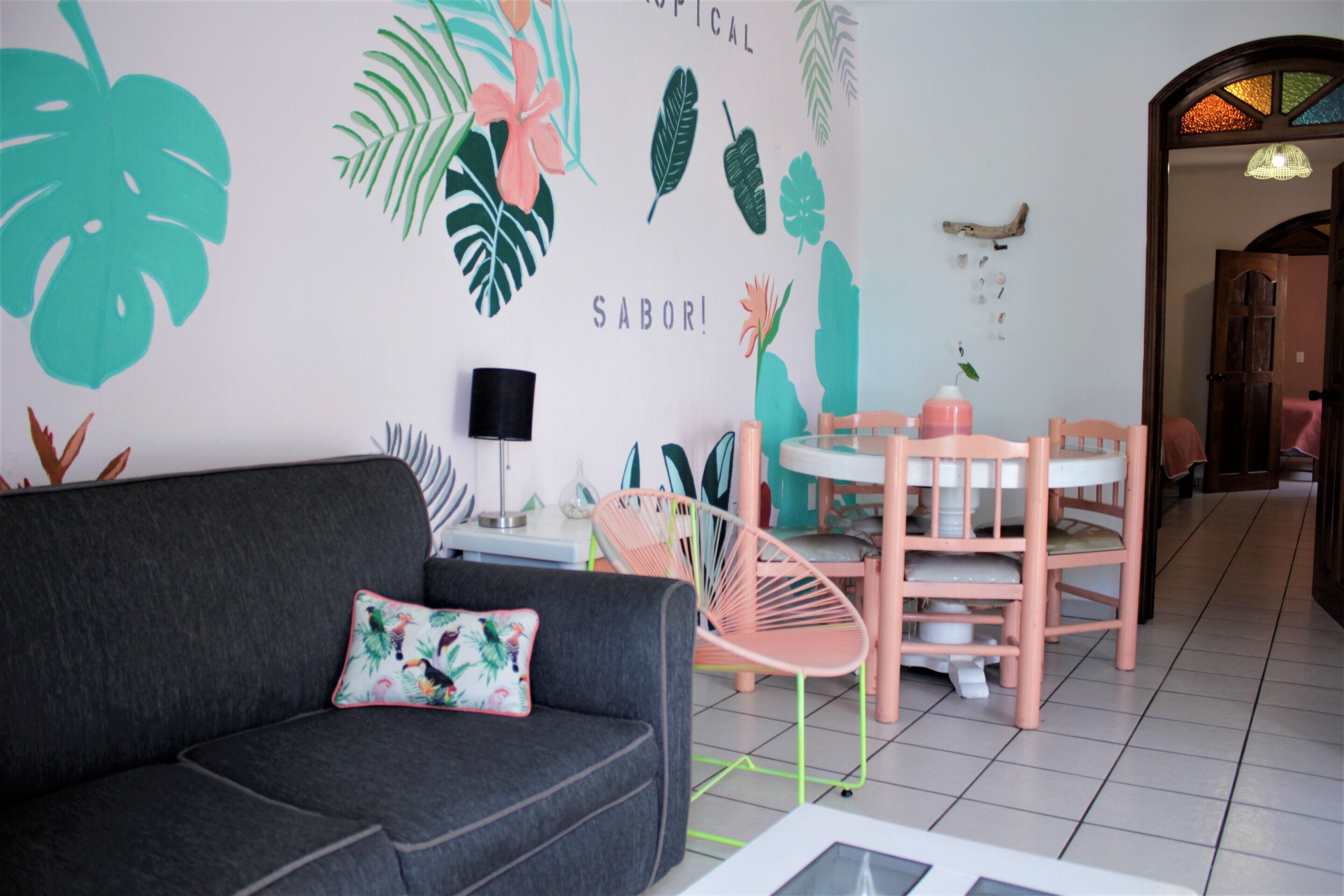 A living room with a grey sofa, a pink chair, a small table, and a wall decorated with tropical leaf patterns and the word "SABOR!".