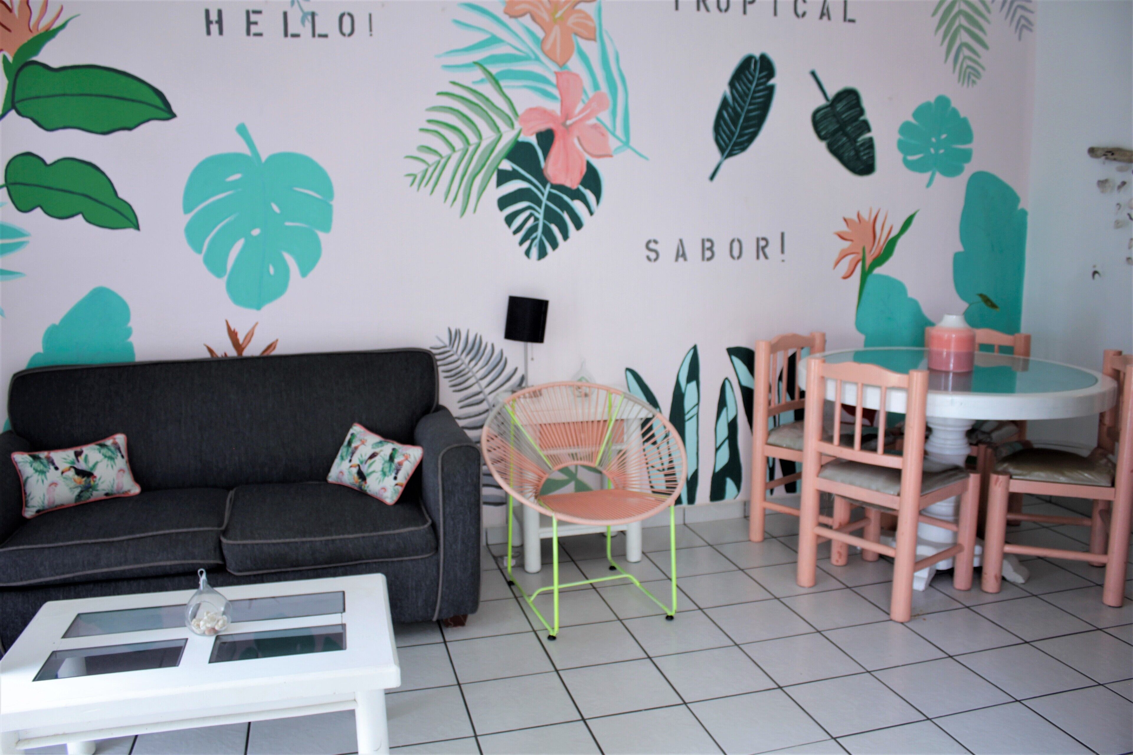 A room with a grey sofa, a white coffee table, a pink chair, and a round table with pink chairs. The wall has tropical leaf patterns and the words "HELLO," "TROPICAL," and "SABOR!" visible.