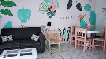 A room with a grey sofa, a white coffee table, a pink chair, and a round table with pink chairs. The wall has tropical leaf patterns and the words "HELLO," "TROPICAL," and "SABOR!" visible.