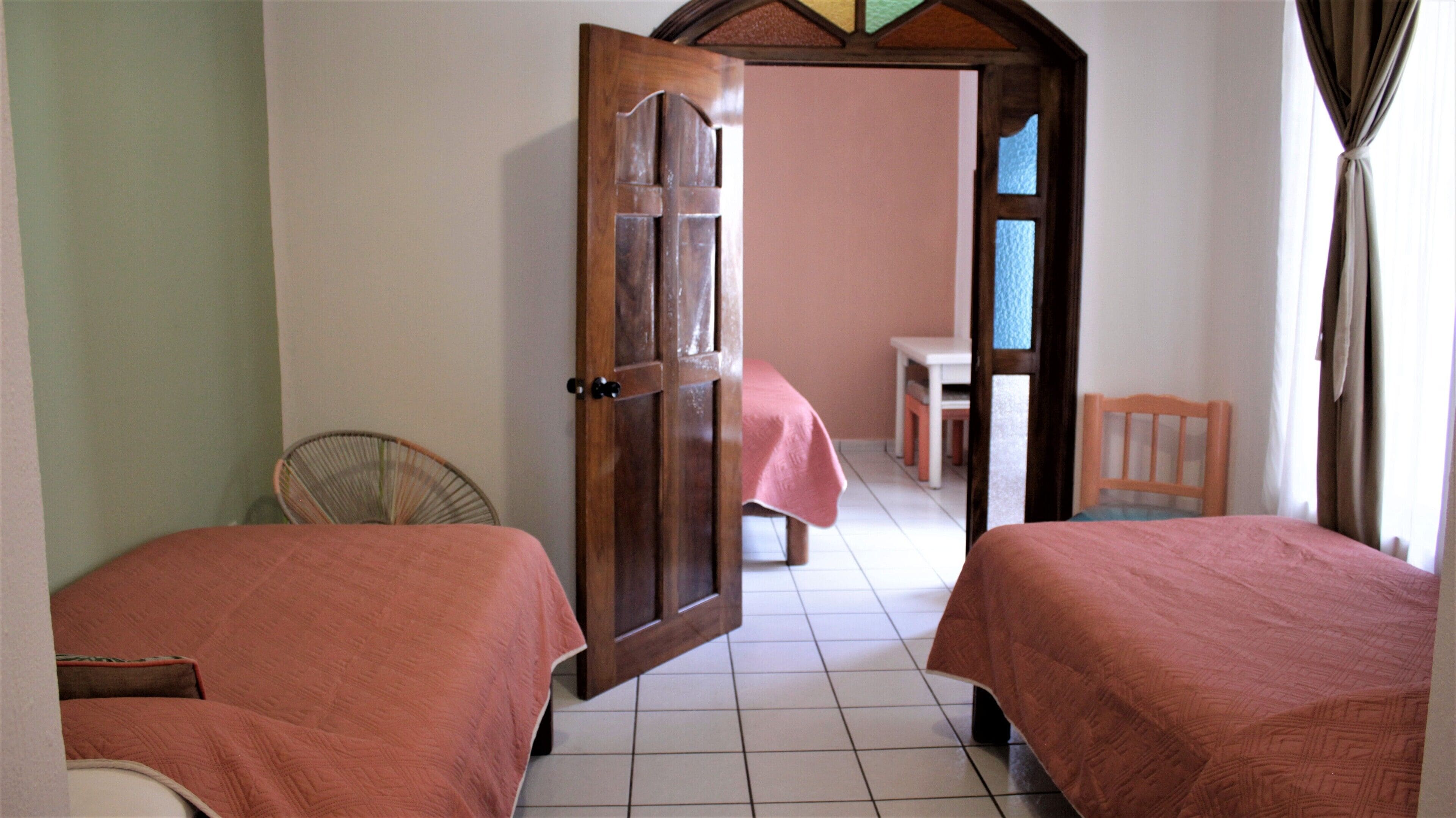 Una habitación con dos camas, una puerta de madera, una silla y una mesita con mantel rosa.