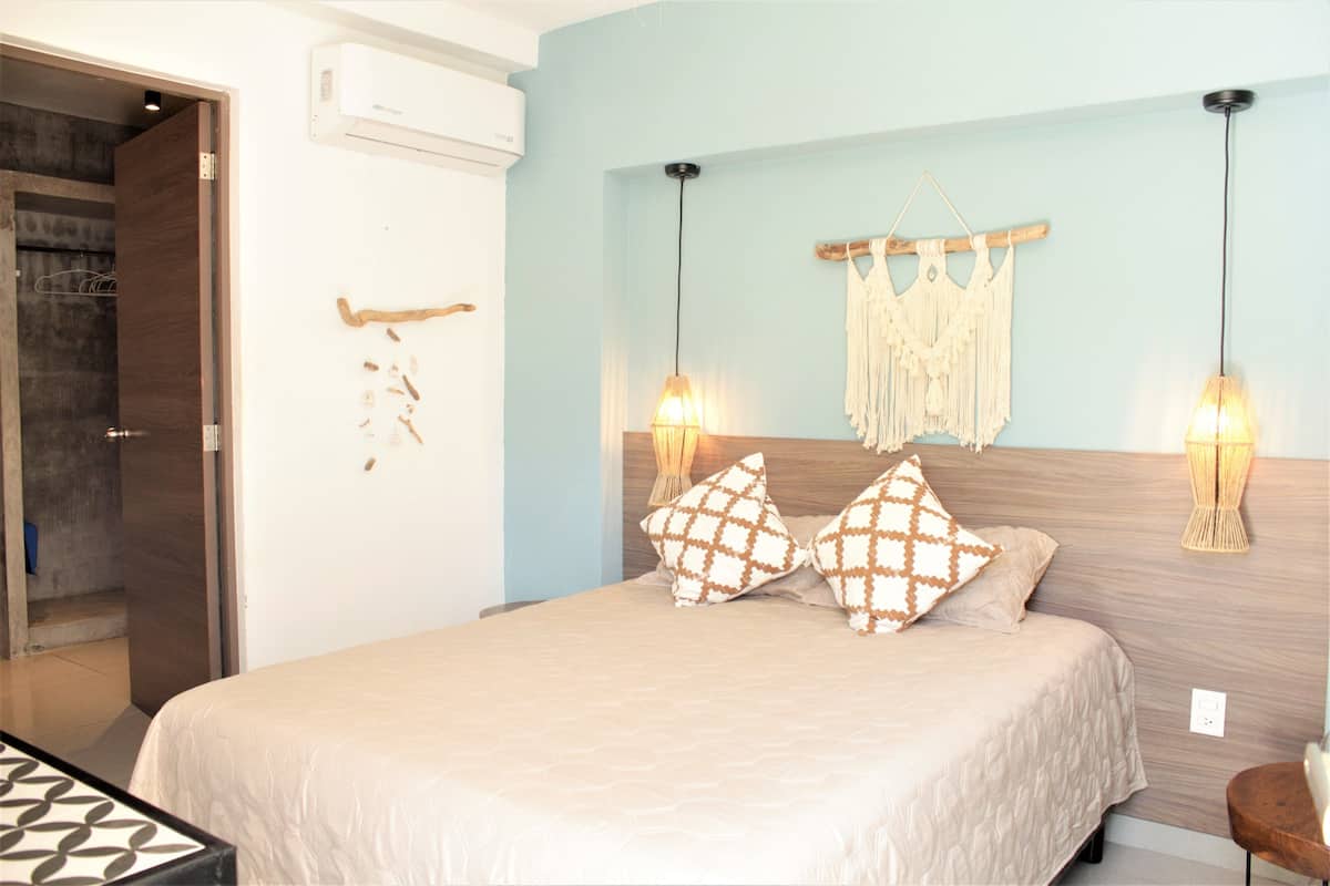Een slaapkamer met een bed, twee wandlampen, een macramé wanddecoratie en een houten deur.