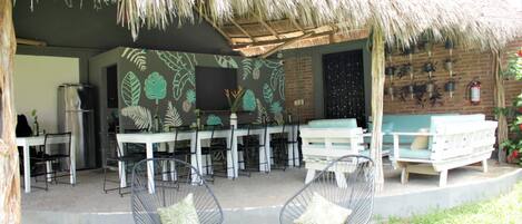An outdoor dining area with a thatched roof, tables, chairs, and a brick wall with potted plants.