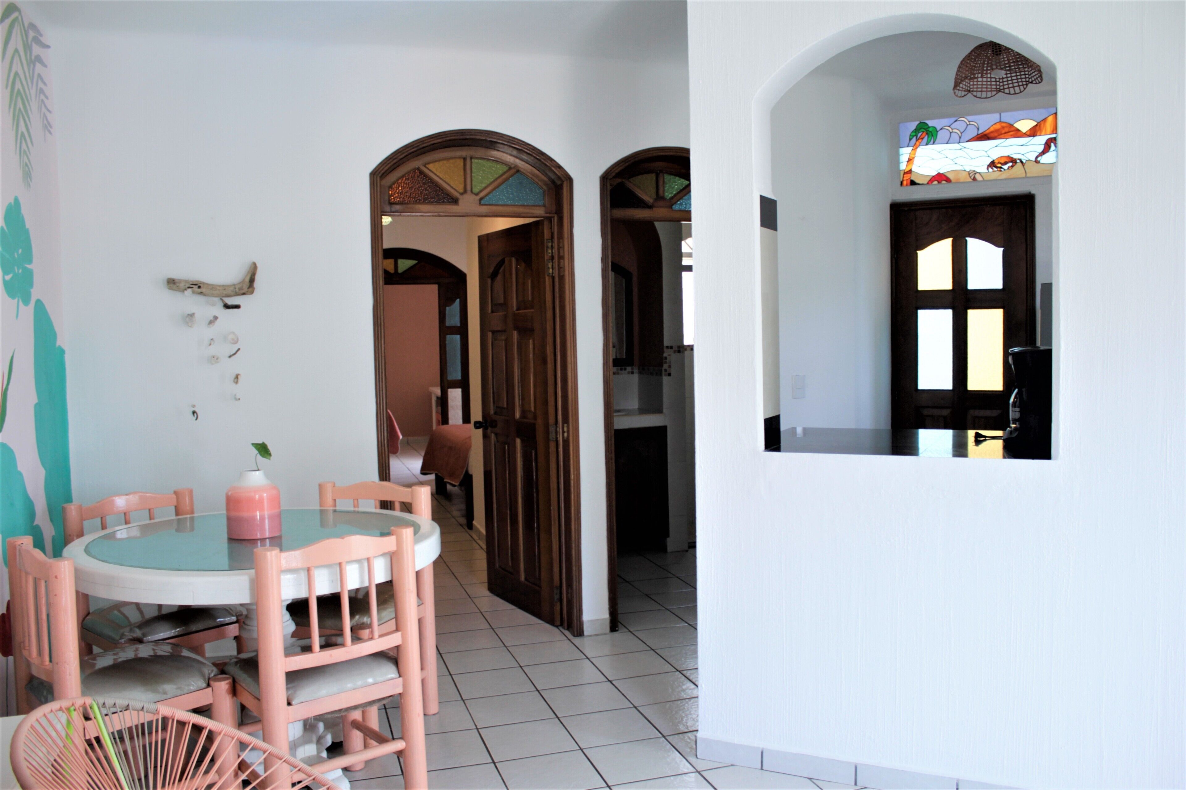 Un comedor con una mesa redonda y sillas, una cocina visible a través de un vano abierto, y una ventana pequeña con vistas al exterior.