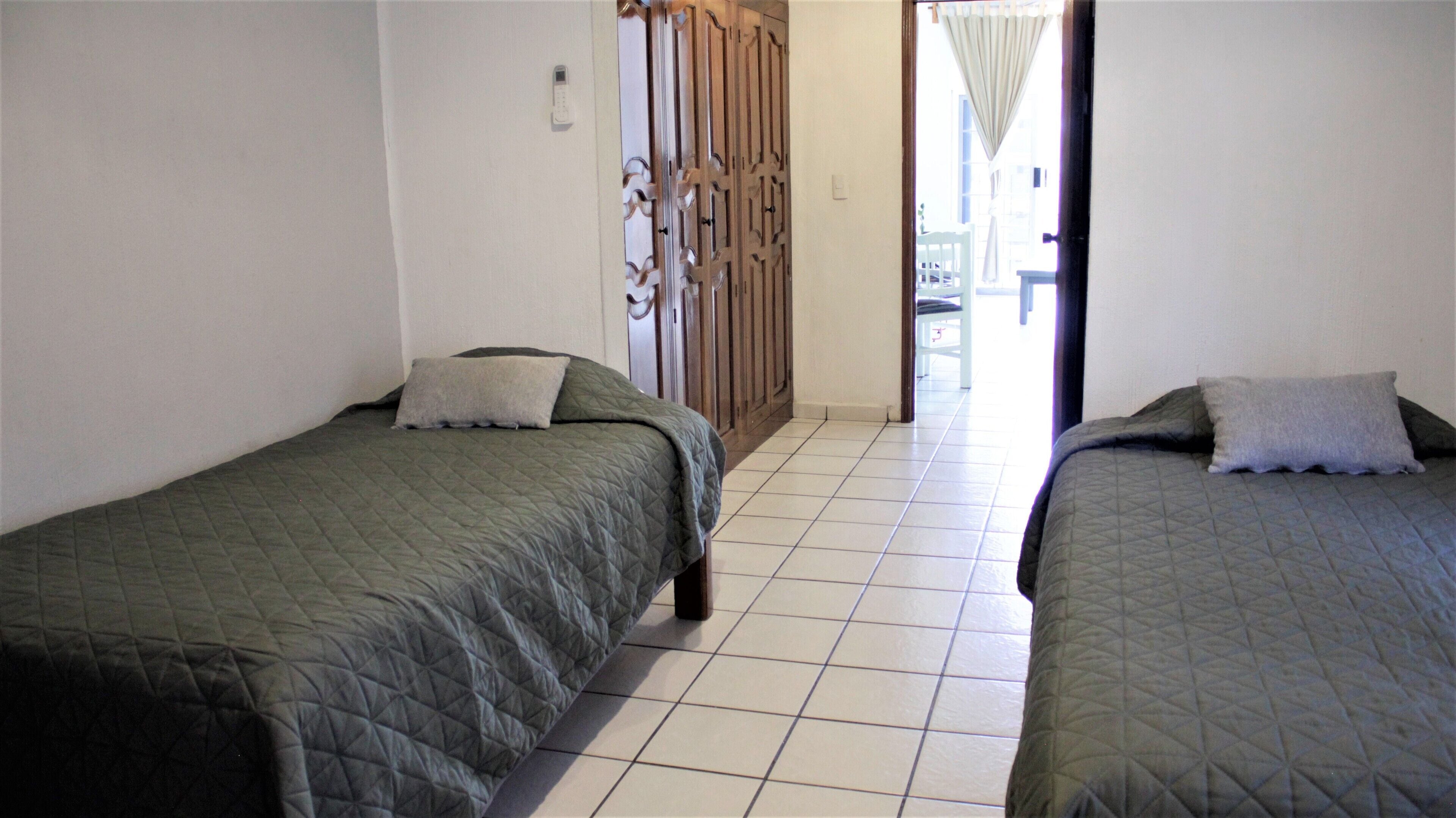 Two single beds with matching green quilts and pillows, a tiled floor, and a wooden wardrobe with a decorative pattern.