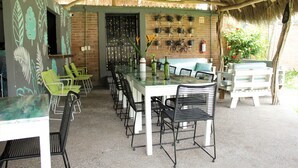 Un espacio de comedor al aire libre con techo de paja, pared de ladrillo con macetas colgantes y diversos sillones y mesas.