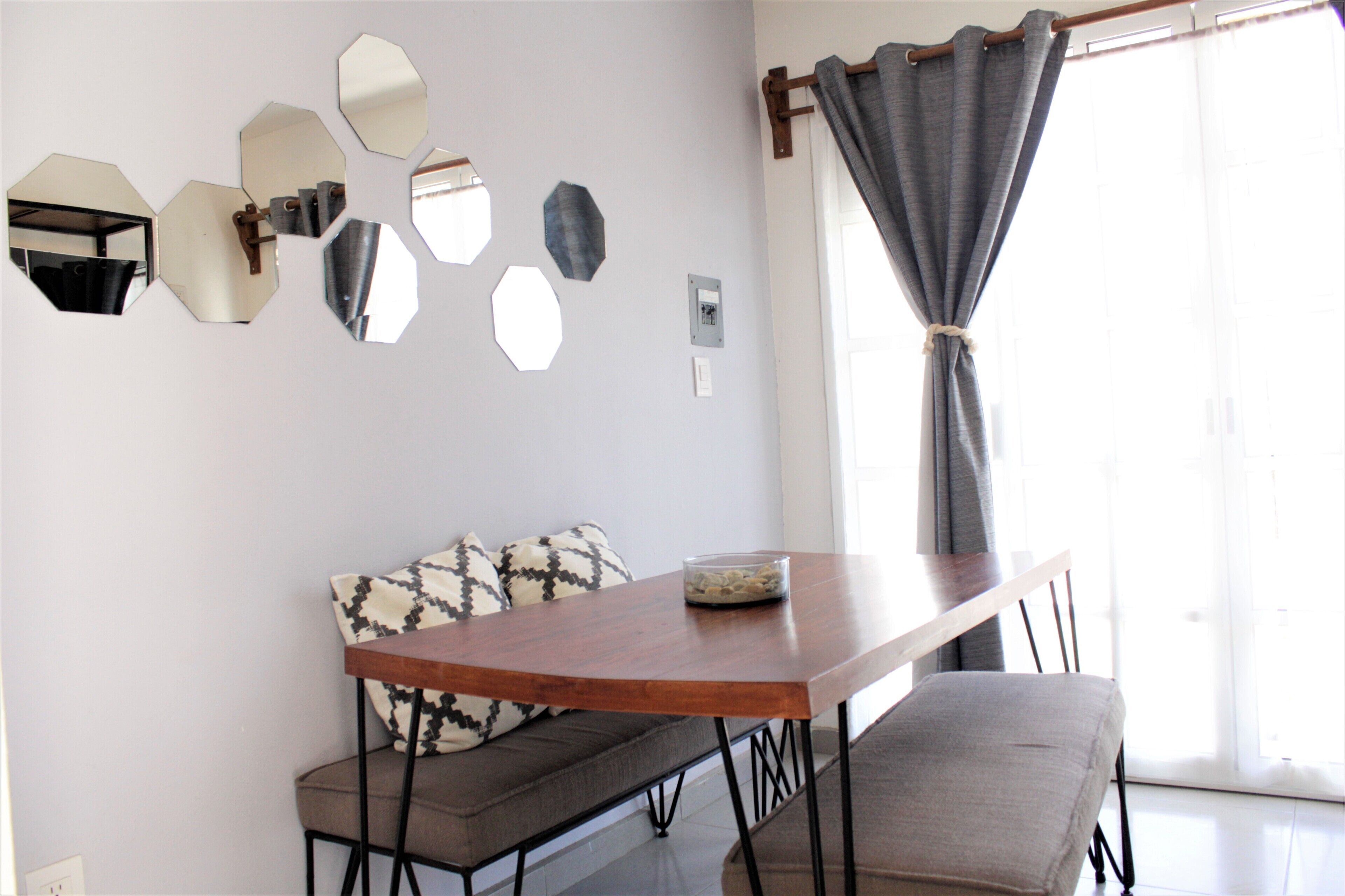 A dining area with a wooden table, a bench, and a sofa. There are multiple wall-mounted mirrors and a window with curtains.
