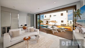 Una sala de estar moderna con un sofá blanco, un ventanal de vidrio que da a una zona de piscina y vistas a un edificio blanco con piscina.