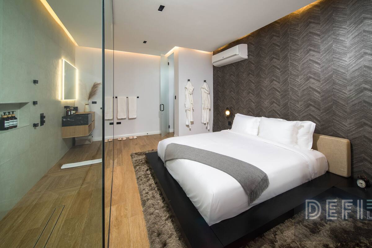 A modern hotel room with a large bed, a wooden floor, a mirror, a vanity with toiletries, and a herringbone-patterned wall.