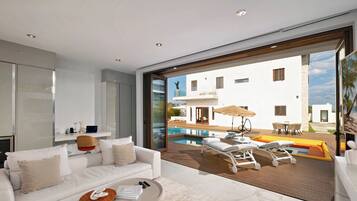 Una sala de estar moderna con un sofá blanco, un ventanal de vidrio que da a una zona de piscina y vistas a un edificio blanco con piscina.