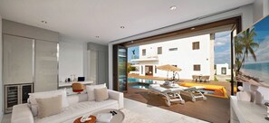 Una sala de estar moderna con un sofá blanco, un ventanal de vidrio que da a una zona de piscina y vistas a un edificio blanco con piscina.