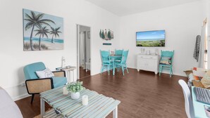 Un salon comprenant un coin repas, agrémenté d’un tableau représentant des palmiers, d’un téléviseur et offrant une vue sur l’océan.