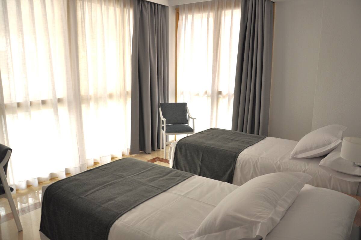 Quarto de hotel com duas camas, uma cadeira e uma janela grande com cortinas.