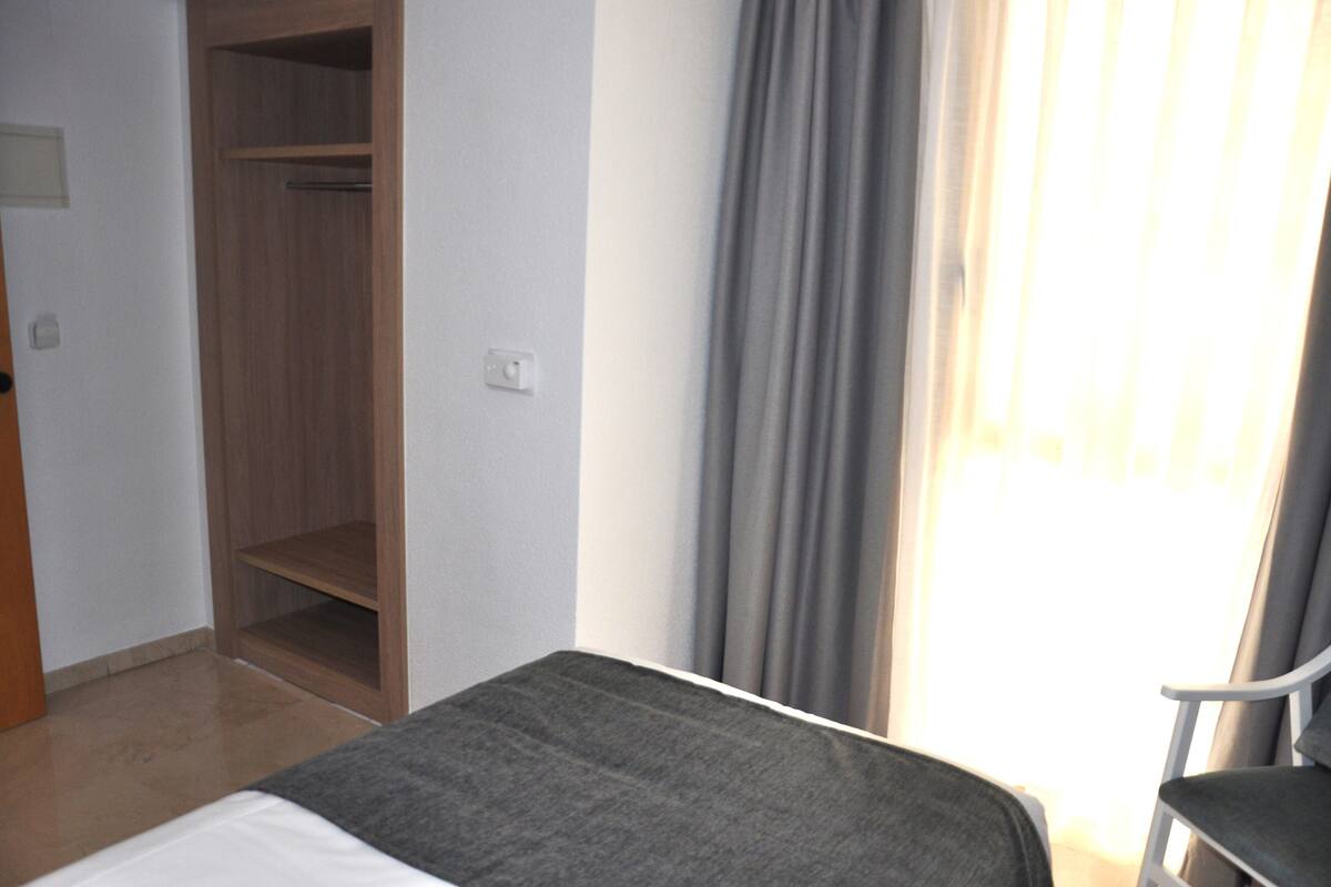 Quarto de hotel com cama, cadeira, guarda-roupa e janela com cortinas.