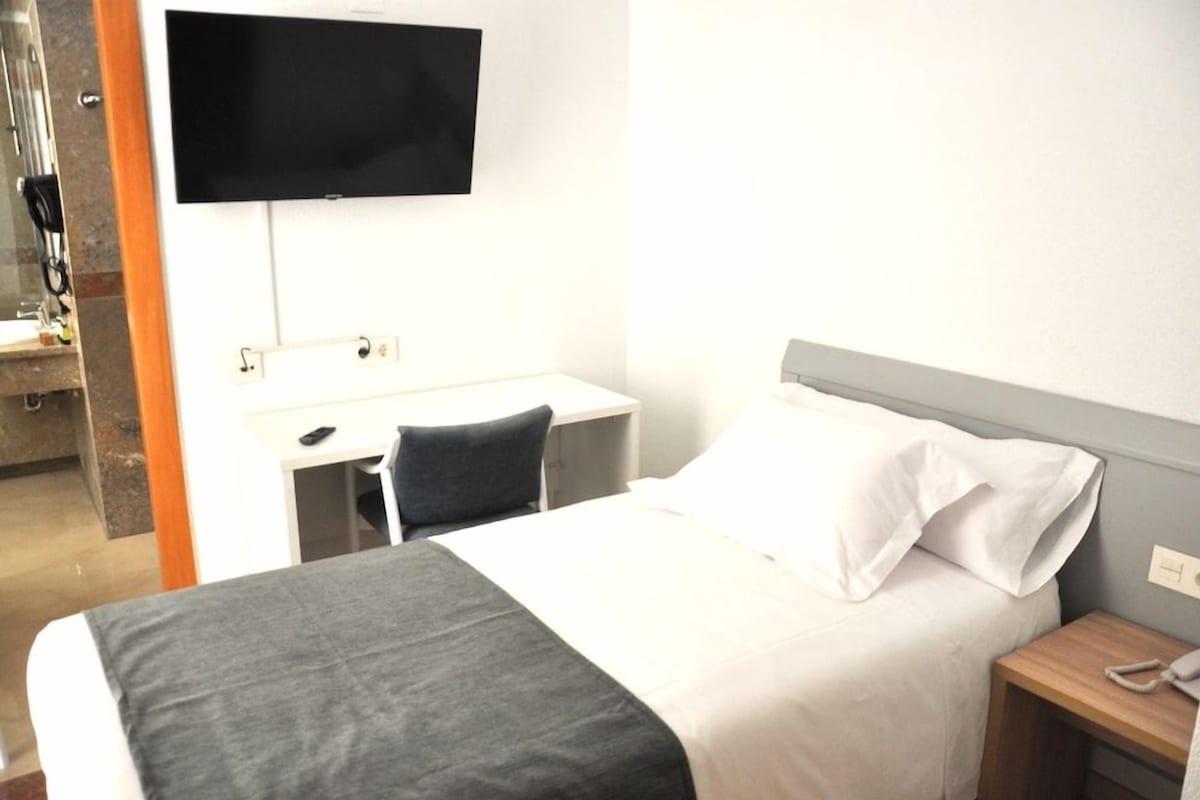 A hotel room with a bed, a desk, a chair, a television mounted on the wall, and a bedside table with a phone.